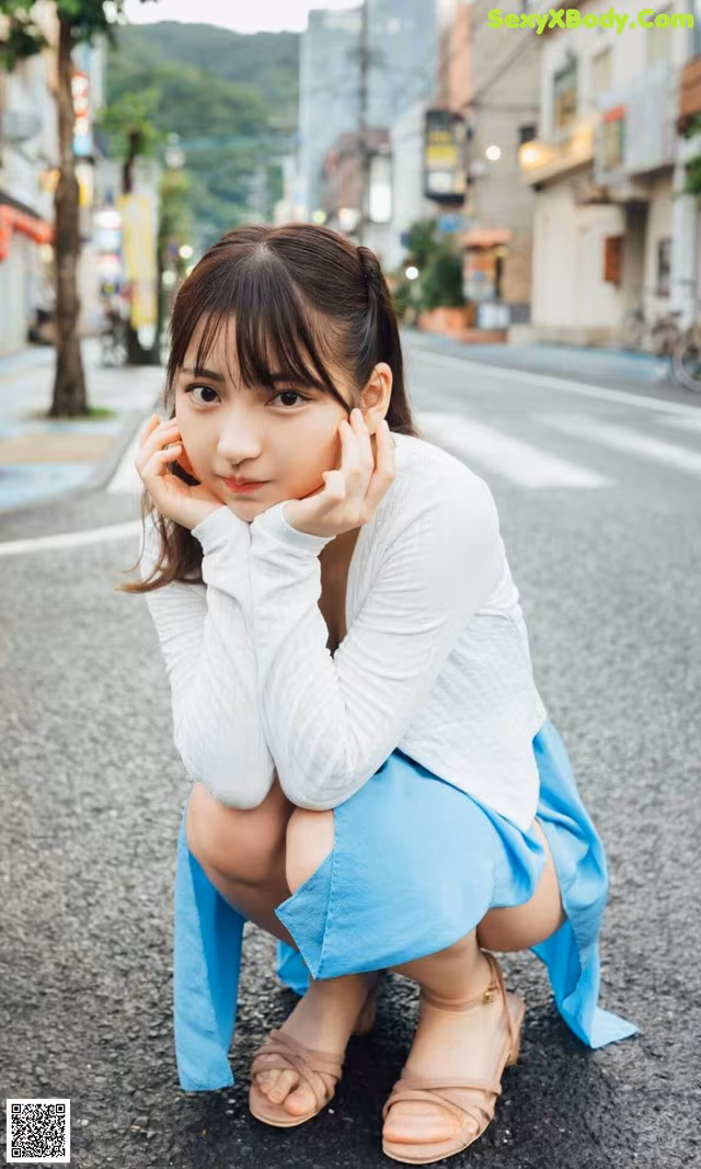 A young woman crouching down on the street with her hand on her chin.