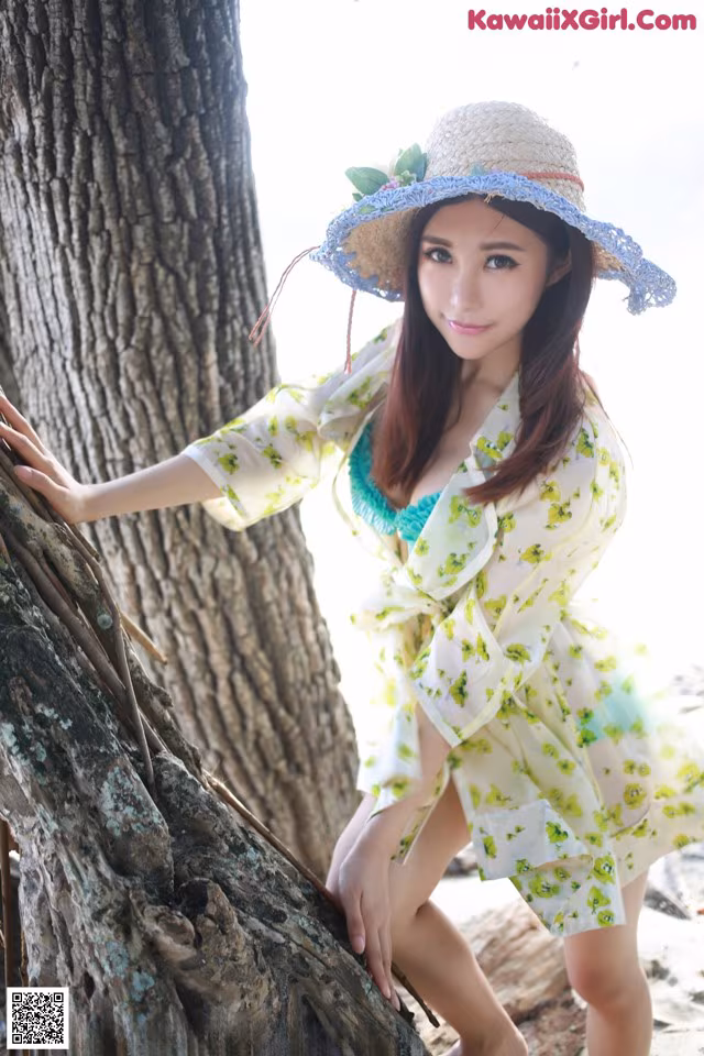 A woman in a yellow dress and a straw hat leaning against a tree.
